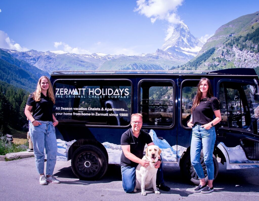 The Zermatt Holidays Team, Cor Groenenboom, Rianne and Jaqueline standing in front of the Zermatt Holidays electro taxi, with Nala the office dog. Zermatt and the Matterhorn in the Background